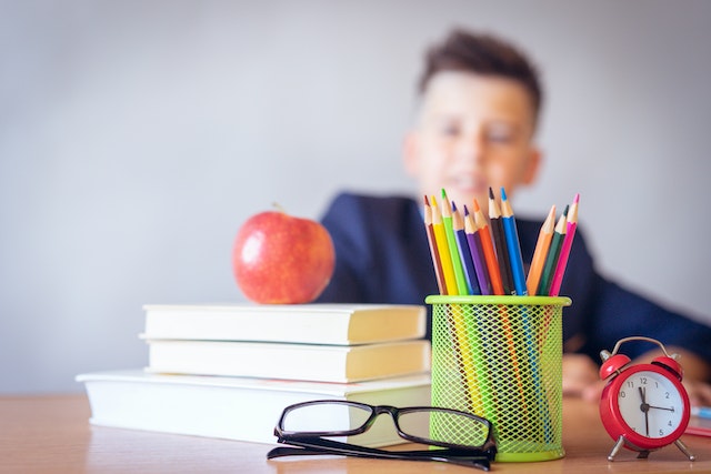 jongen zit aan aan een schooltafel met een appel, bril en potloden
